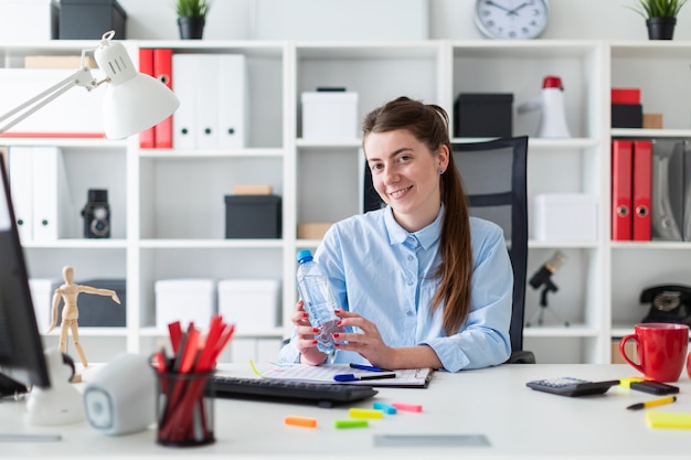 a young girl sits at a table in the office and holds a bottle of water in her hand.