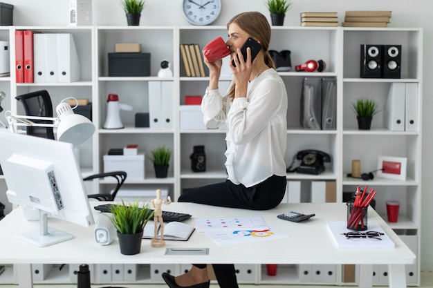 Foto ragazza che si siede allo scrittorio in ufficio, tenendo una tazza rossa in sua mano e parlando sul telefono