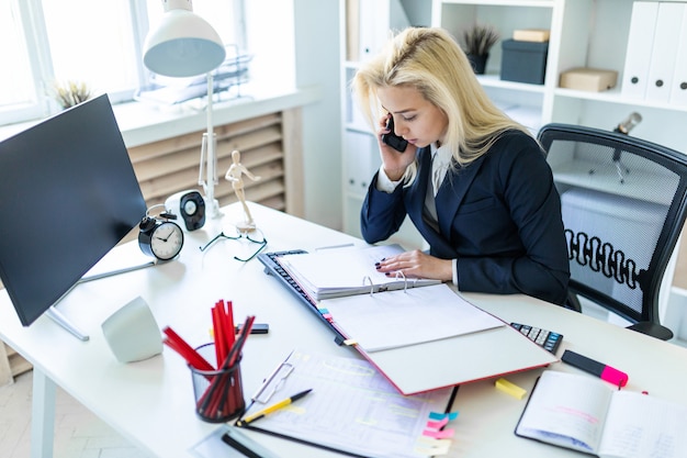 Young girl sitting at desk in office, talking on phone and looking at documents.