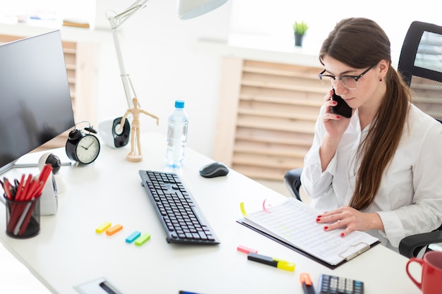 Young girl sitting at desk in office, talking on phone and working with documents