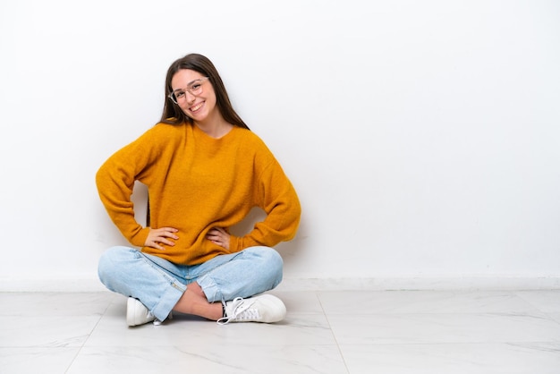 Young girl sitting on the floor isolated on white background posing with arms at hip and smiling