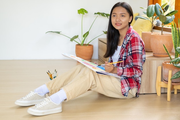 Молодая девушка сидит на полу и рисует на бумаге дома Хобби и изучение искусства дома