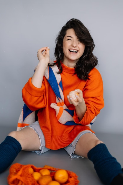 Young girl sitting on the floor in a photo studio with tangerines in her hands posing for a photo throwing tangerines into the camera laughing and having fun Bright emotions