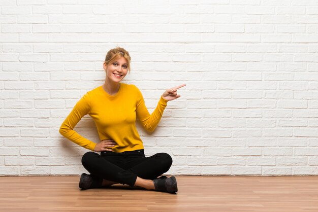 Young girl sitting on the floor pointing finger to the side and presenting a product