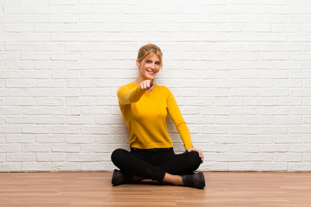 Young girl sitting on the floor points finger at you with a confident expression