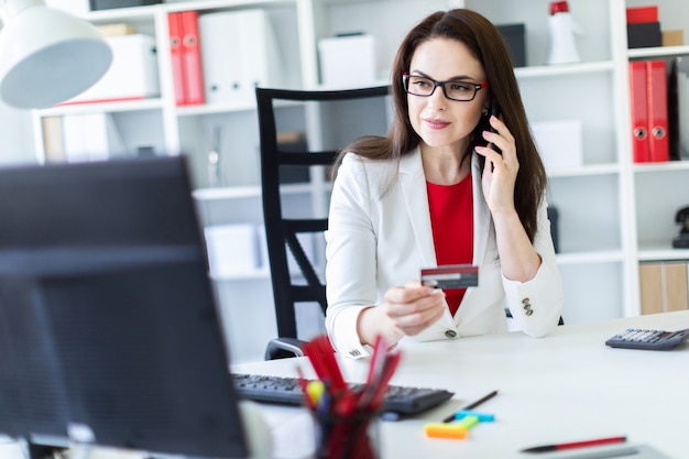 A young girl sitting in the office at the table and holding a Bank card and phone.