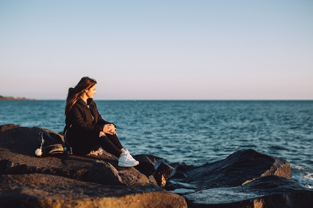 Young girl sitting on the stones by the sea and looks at the sunset.