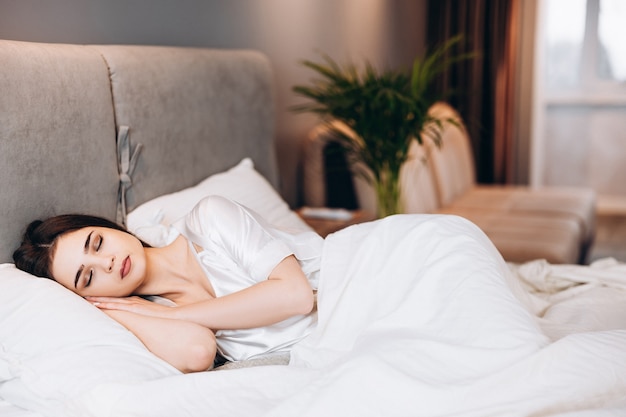 Young girl sleeping in bed. Morning or evening in bed with the female model. Brunette in silk pajamas with closed eyes in the bed in the bedroom.