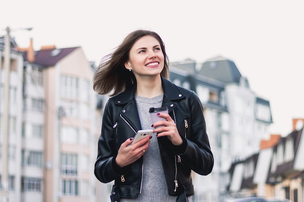 A young girl smiles and walks around the city with a mobile phone and a cup of coffee