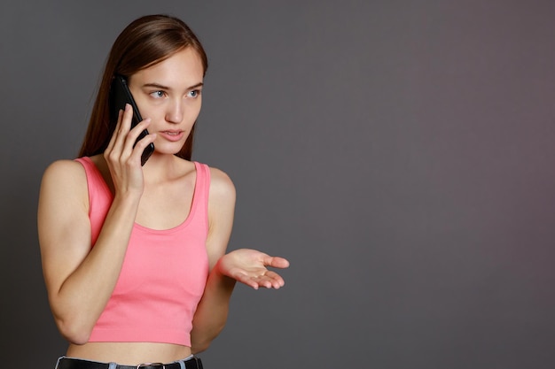 Молодая девушка стоит на сером фоне и разговаривает по телефону