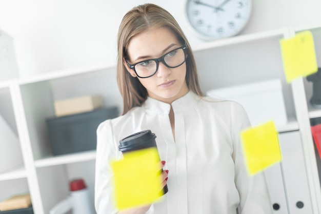 Una giovane ragazza in piedi vicino al consiglio con adesivi in ufficio. nelle mani della ragazza un bicchiere di caffè.