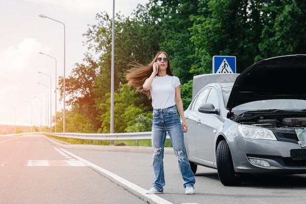 A young girl stands near a broken car in the middle of the highway and calls for help on the phone, while trying to stop passing cars. Failure and breakdown of the car. Waiting for help.