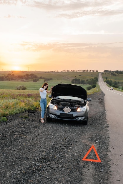 A young girl stands near a broken car in the middle of the highway during sunset and tries to call for help on the phone and start the car Waiting for help Car service Car breakdown on the road