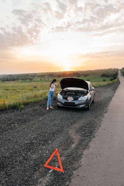 A young girl stands near a broken car in the middle of the highway during sunset and tries to call for help on the phone and start the car Waiting for help Car service Car breakdown on the road