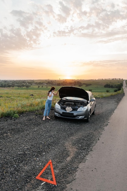 Молодая девушка стоит возле сломанной машины посреди шоссе во время заката и пытается позвать на помощь по телефону и завести машину В ожидании помощи Автосервис Поломка машины на дороге