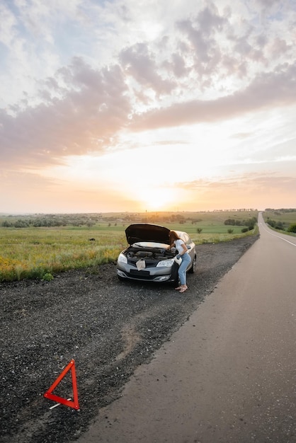 A young girl stands near a broken car in the middle of the highway during sunset and tries to repair it. Breakdown and repair of the car. Troubleshooting the problem.