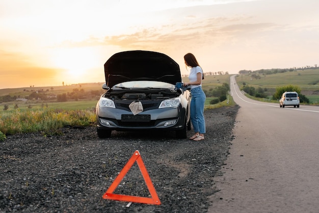 若い女の子が日没時に高速道路の真ん中で壊れた車の近くに立って、それを修理しようとします問題のトラブルシューティング助けを待っています車のサービスroadxAでの車の故障