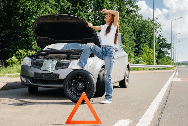 高速道路の真ん中で壊れた車の近くに立っている若い女の子が、暑い晴れた日に壊れたホイールを交換しようとします。車の故障と故障。助けを待っています。