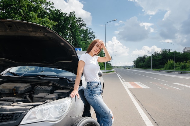 若い女の子が高速道路の真ん中で壊れた車輪で壊れた車の近くに立って、暑い日に助けを待ってイライラしています。車の故障と故障。助けを待っています。