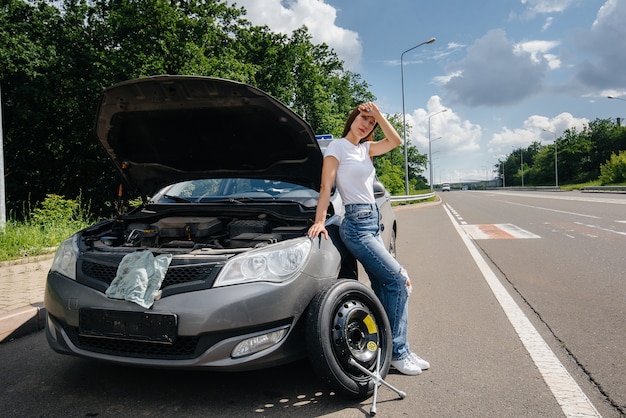 A young girl stands near a broken car with a broken wheel in the middle of the highway and is frustrated waiting for help on a hot day. Breakdown and breakdown of the car. Waiting for help.