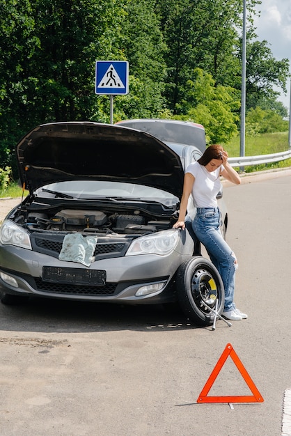 A young girl stands near a broken car with a broken wheel in the middle of the highway and is frustrated waiting for help on a hot day. Breakdown and breakdown of the car. Waiting for help.