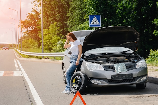 若い女の子は、高速道路の真ん中で壊れたホイールで壊れた車の近くに立って、暑い日に助けを待ってイライラしています