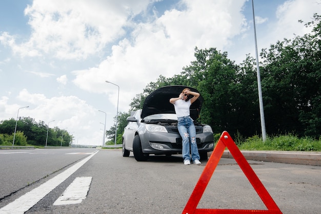 A young girl stands near a broken-down car in the middle of the highway and calls for help on the phone. Failure and breakdown of the car. Waiting for help.