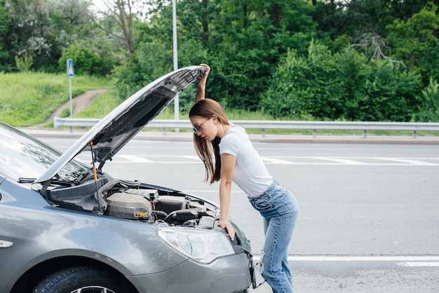 A young girl stands near a broken-down car in the middle of the highway and looks under the hood. Failure and breakdown of the car. Waiting for help.