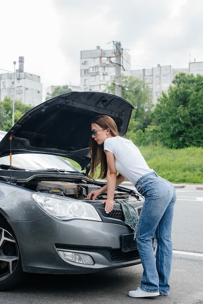 A young girl stands near a broken-down car in the middle of the highway and looks under the hood. Failure and breakdown of the car. Waiting for help.