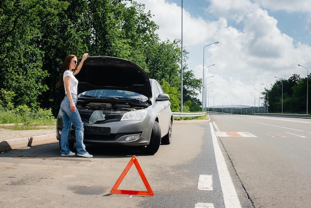 高速道路の真ん中で壊れた車の近くに立っている若い女の子が、ボンネットの下を見ています。車の故障と故障。助けを待っています。