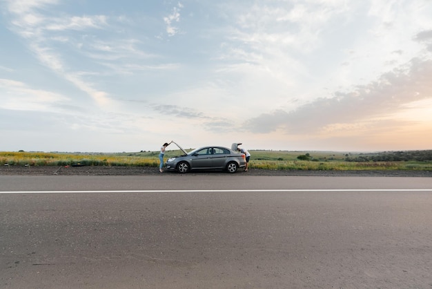 A young girl stands near a brokendown car in the middle of the highway during sunset and tries to call for help on the phone Waiting for help Car service Car breakdown on road