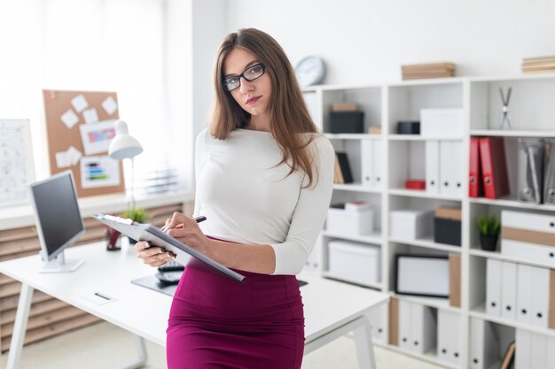 A young girl stands near a computer Desk and holds a tablet with sheets for records.