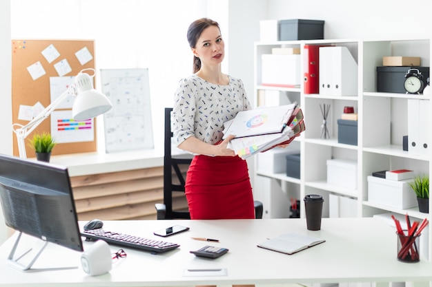 A young girl stands near a computer Desk in the office and holds folders and graphics.