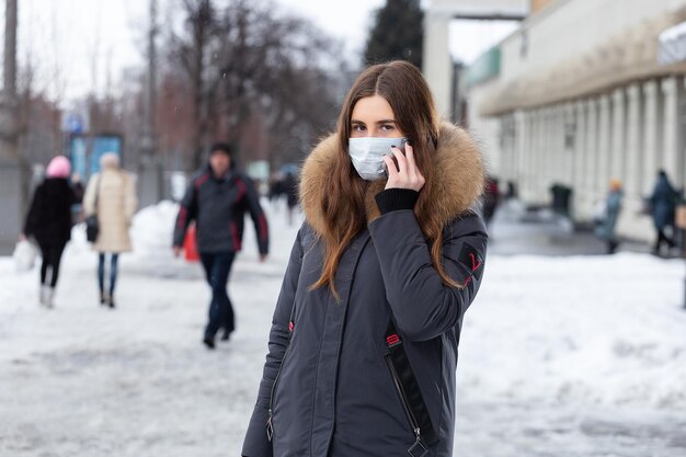 A young girl stands near the road and talking on the smartphone in a medical mask Protection against virus outbreaks flu and air pollution from cars in the city