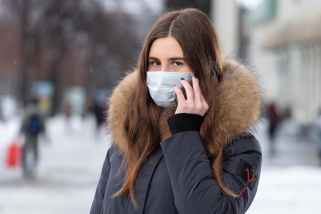 若い女の子が道路の近くに立って、医療用マスクでスマートフォンで話している市内の車からのウイルス発生インフルエンザと大気汚染に対する保護