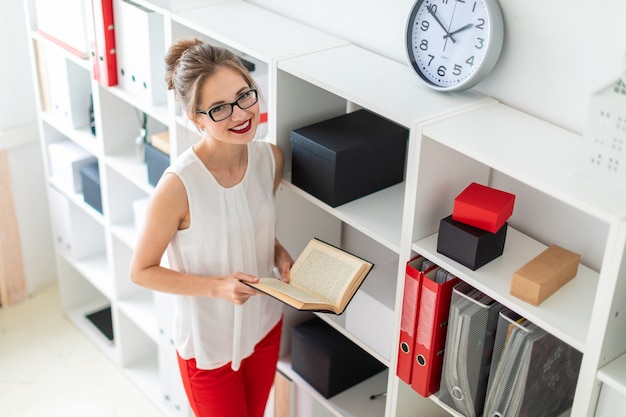 A young girl stands near the shelf and holds an open book in her hands.