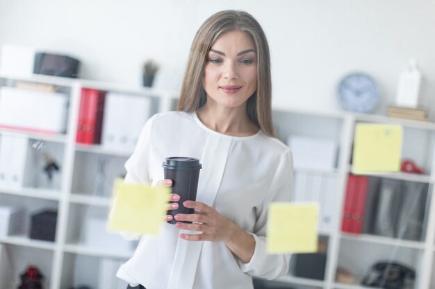 若い女の子がステッカー付きの透明なボードの近くに立って、コーヒーを1杯持っています。