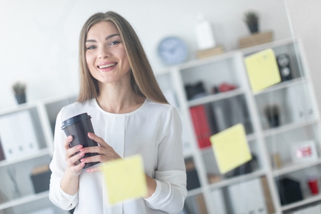 若い女の子がステッカー付きの透明なボードの近くに立って、コーヒーを1杯持っています。