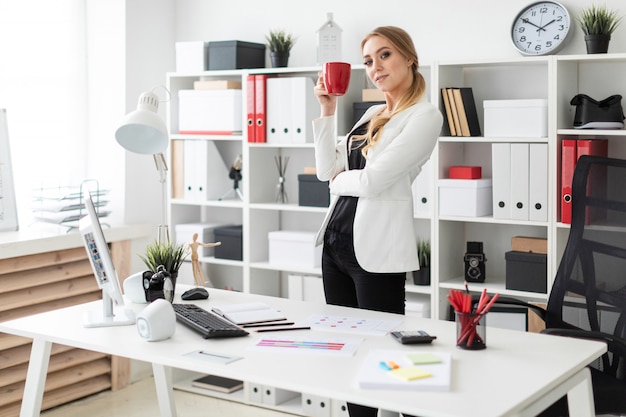 若い女の子がコンピューターの机の近くのオフィスに立ち、彼女の手に赤いカップを持っています。