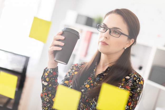 A young girl stands in the office near a transparent Board with stickers and drinks coffee.