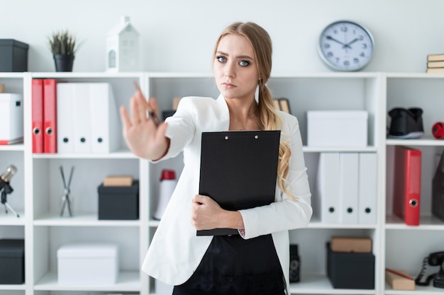 A young girl stands in the office next to the rack, holds documents and a pen in her hand and shows a stop sign.