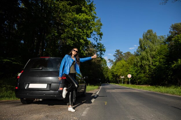 Photo a young girl stands on the road in a green forest next to a broken car and asks for help stopping other cars