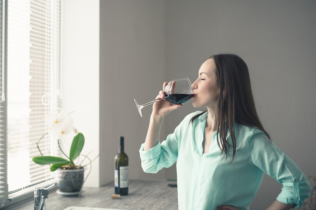 A young girl stands at the window in the kitchen and drinks from a glass of red wine