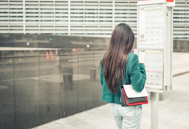 A young girl stands with her back near a bus stop with a timetable