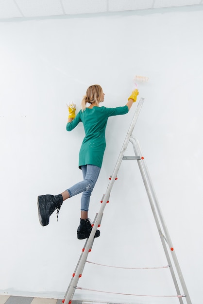 A young girl on a stepladder paints a white wall with a roller. Repair of the interior.