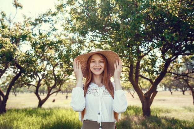 Young girl in a straw hat travels green meadows. Beautiful red-haired hipster woman in a green garden