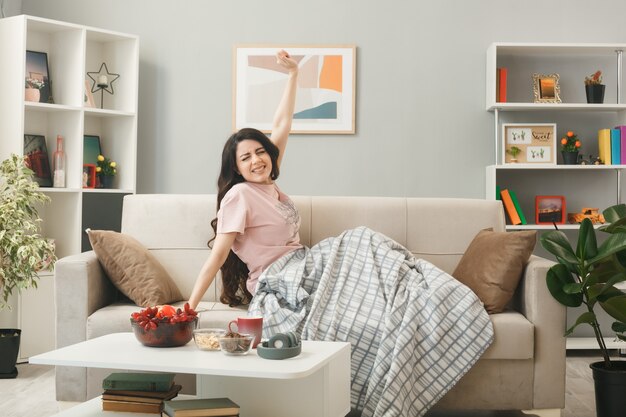 Young girl stretching out arm wrapped in plaid sitting on sofa behind coffee table in living room