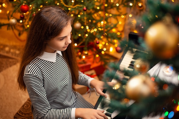 Young girl in striped dress playing chtistmas songs on the piano. festive bright holidays decoration on the background