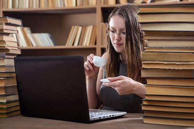 若い女の子の学生は、図書館のラップトップでコーヒーを飲みます