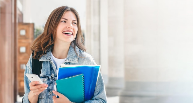 Young girl student laughing against university holds folders notebooks and mobile phone